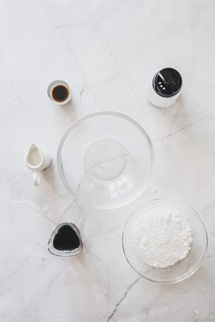 A glass bowl, powdered sugar, milk, vanilla, and two saucesโperfect ingredients for creating delicious Maple Glazed Donutsโare artfully arranged on a white marble surface.