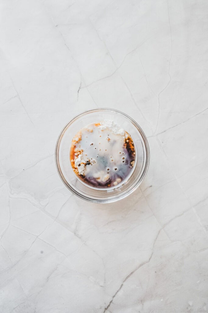 A glass bowl with soy sauce, vinegar, and white powder sits on a white marbled surfaceโits simplicity a striking contrast to the sweet indulgence of maple glazed donuts.
