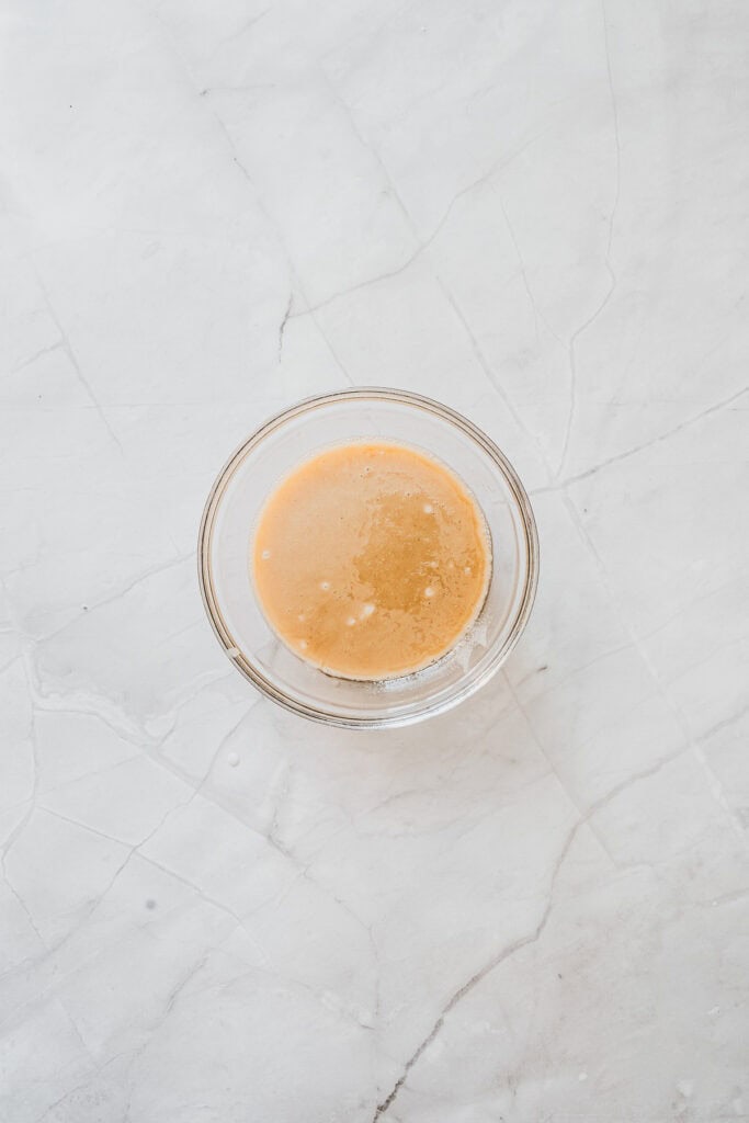 A small glass bowl with a light brown liquid, reminiscent of the glaze used on maple donuts, sits on a white marble surface.