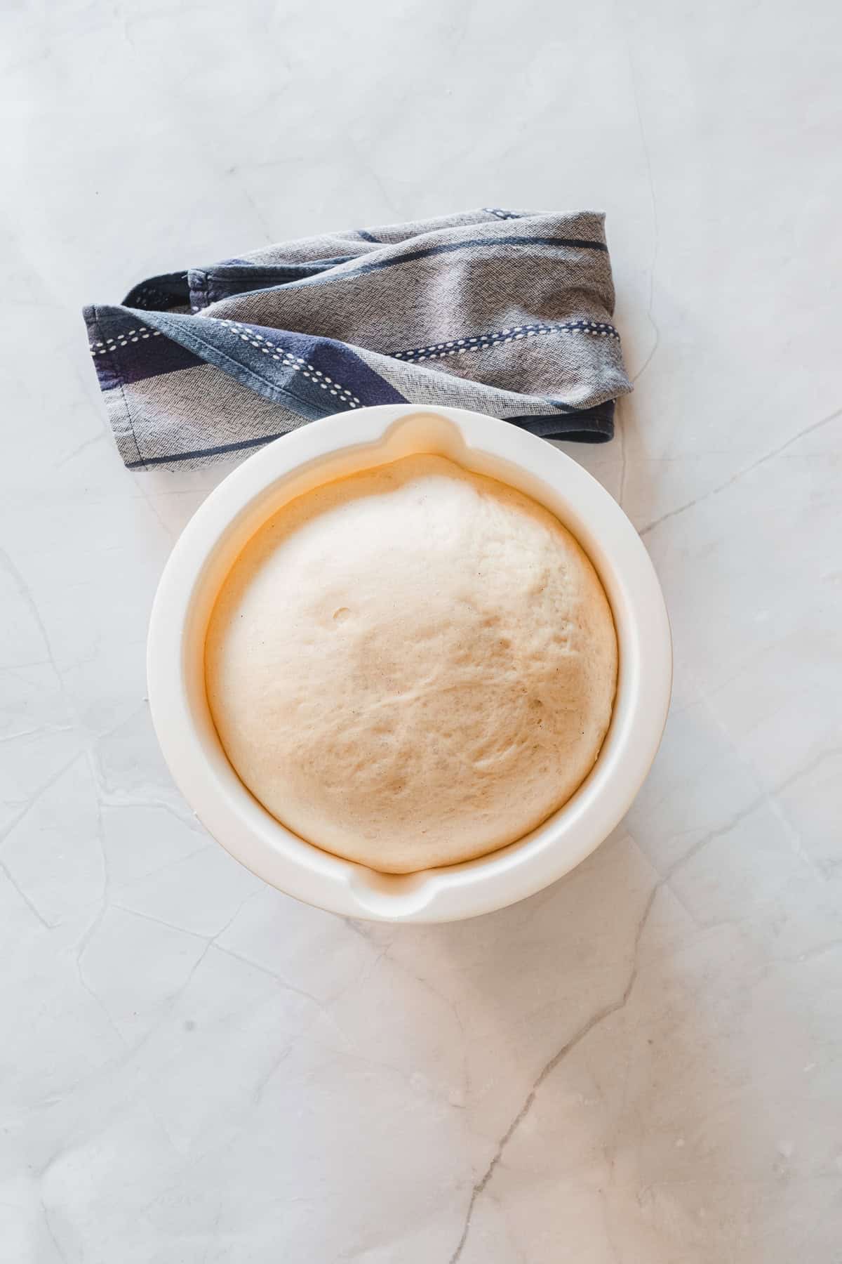 A bowl of risen dough, perfect for making Maple Glazed Donuts, sits next to a folded kitchen towel on a marble countertop.