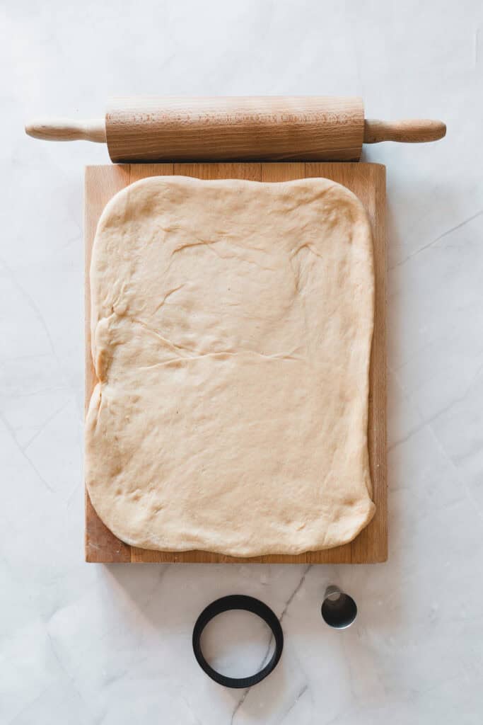 Rectangular pizza dough on a wooden board with a rolling pin, round cutter, and piping tip nearbyโperfect tools not only for crafting delicious pizzas but also for shaping pastries like Glazed Donuts or Maple Glazed Donuts.