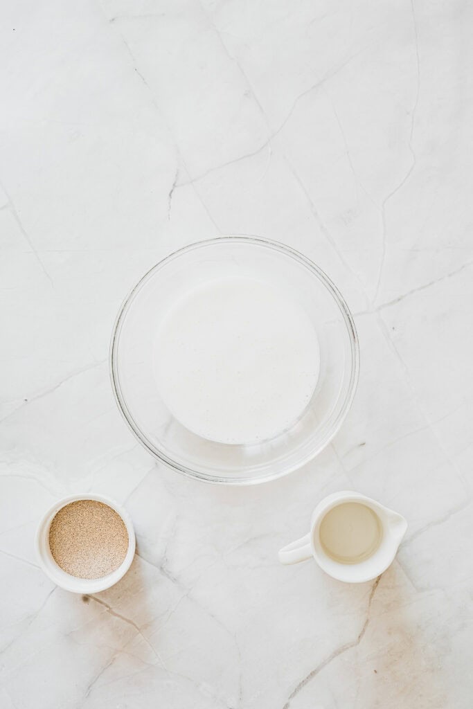 A bowl of foamy liquid, a small cup of yeast, and a small pitcher of liquid on a marble surfaceโperfect ingredients for creating homemade maple glazed donuts.