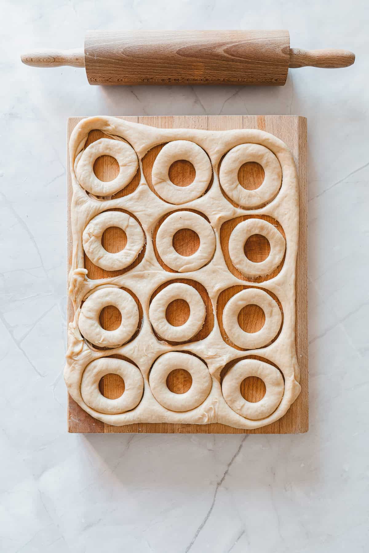 Doughnut shapes cut in dough on a wooden board, rolling pin above, on a white countertopโperfect for crafting homemade Maple Glazed Donuts or classic Glazed Donuts.