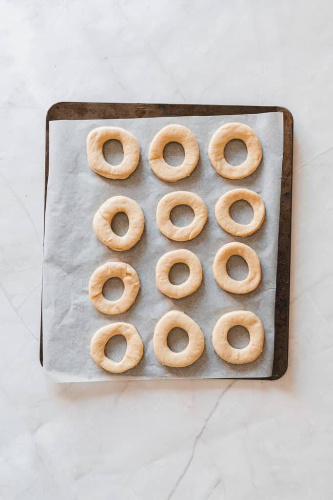 Unbaked doughnut rings, soon to become delicious Maple Glazed Donuts, are arranged in neat rows on a parchment-lined baking tray.