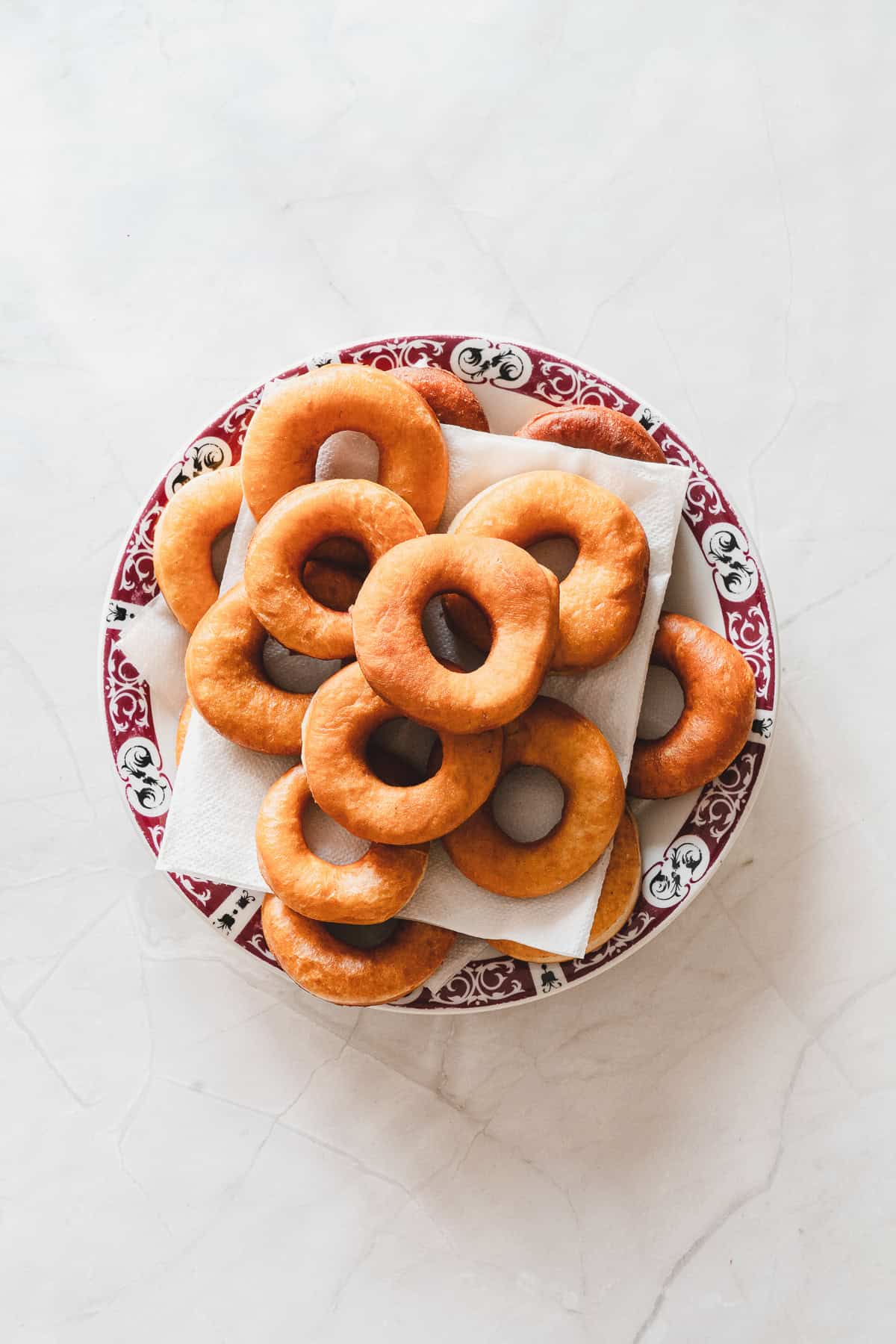 A plate of plain ring doughnuts and maple glazed donuts stacked on napkins, seen from above, on a white surface.