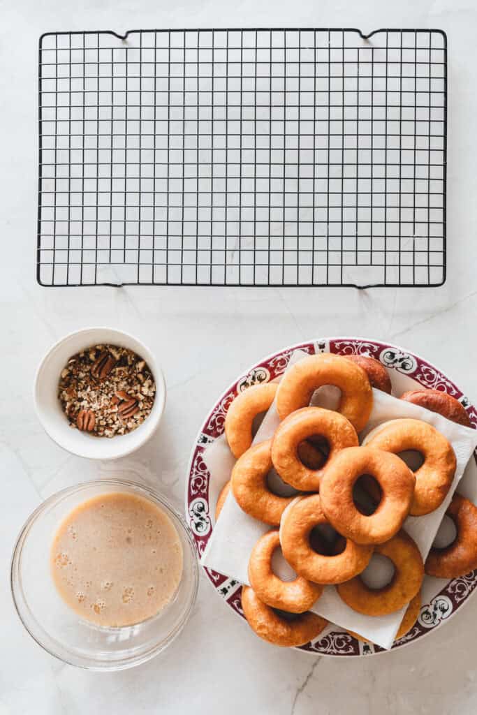 A plate of plain donuts, a bowl of glaze ready for making Glazed Donuts, a bowl of nuts, and an empty cooling rack on a white surface invite you to create delicious Maple Glazed Donuts.