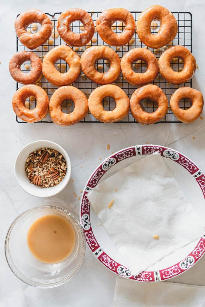 Maple glazed donuts on a cooling rack, with coffee, chopped nuts, and a plate lined with paper towels nearby.