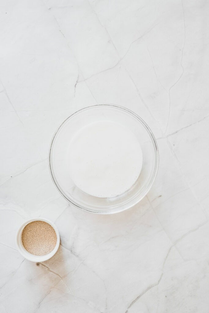 A glass bowl of foamy liquid and a small bowl of granulated yeast sit on a white marble surface, ready to become the perfect base for delicious Maple Glazed Donuts.