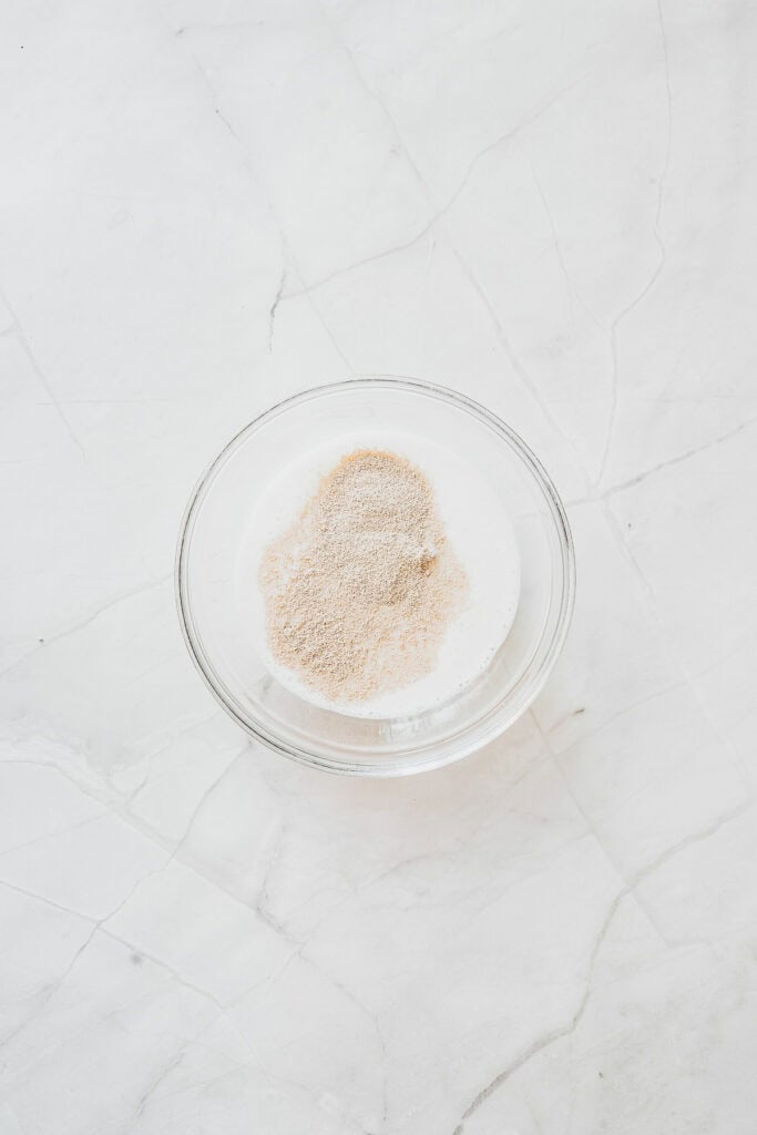 A glass bowl with flour and yeast, ready to become delicious Maple Glazed Donuts, sits on a white marble surface.