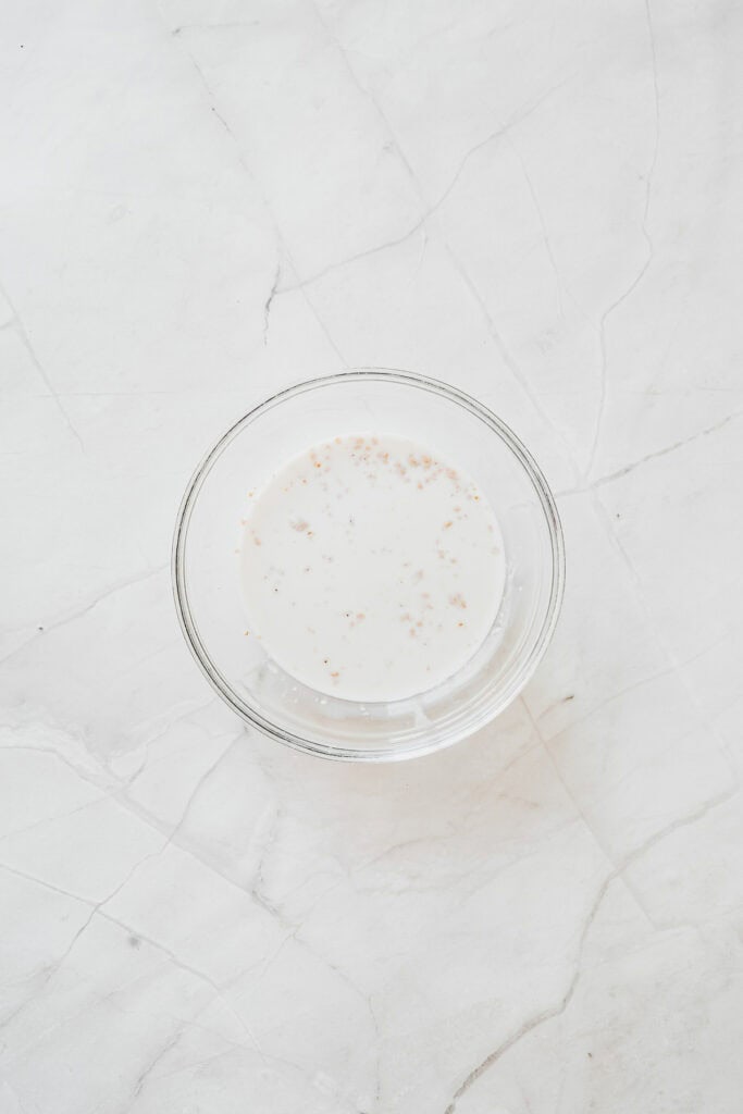 A glass bowl of white liquid with small brown specks on a white marble surface, perfect for glazing Maple Donuts or adding a finishing touch to homemade Glazed Donuts.