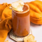 A glass of non-alcoholic apple cider topped with dried apple slices sits beside an orange cloth and more dried apple slices, creating a cozy autumn display.
