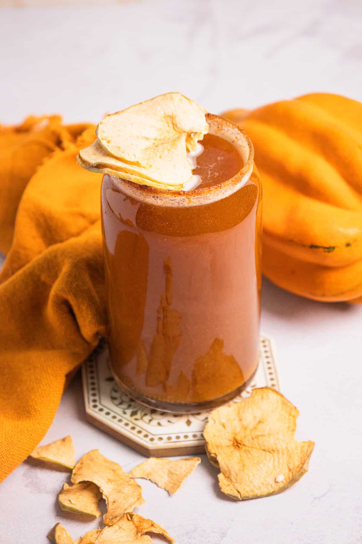 A glass of non-alcoholic apple cider topped with dried apple slices sits beside an orange cloth and more dried apple slices, creating a cozy autumn display.