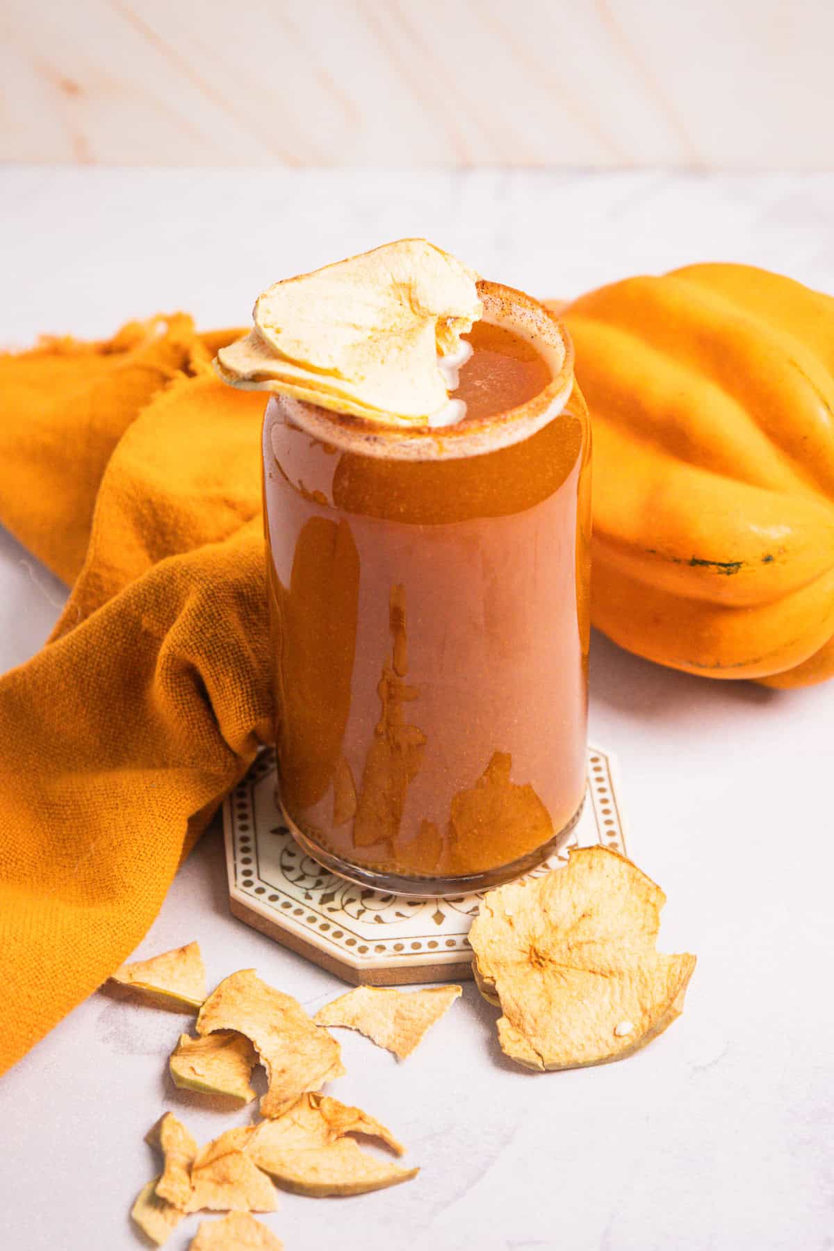 A glass of non-alcoholic pumpkin smoothie topped with apple chips, surrounded by dried apple slices and a pumpkin.