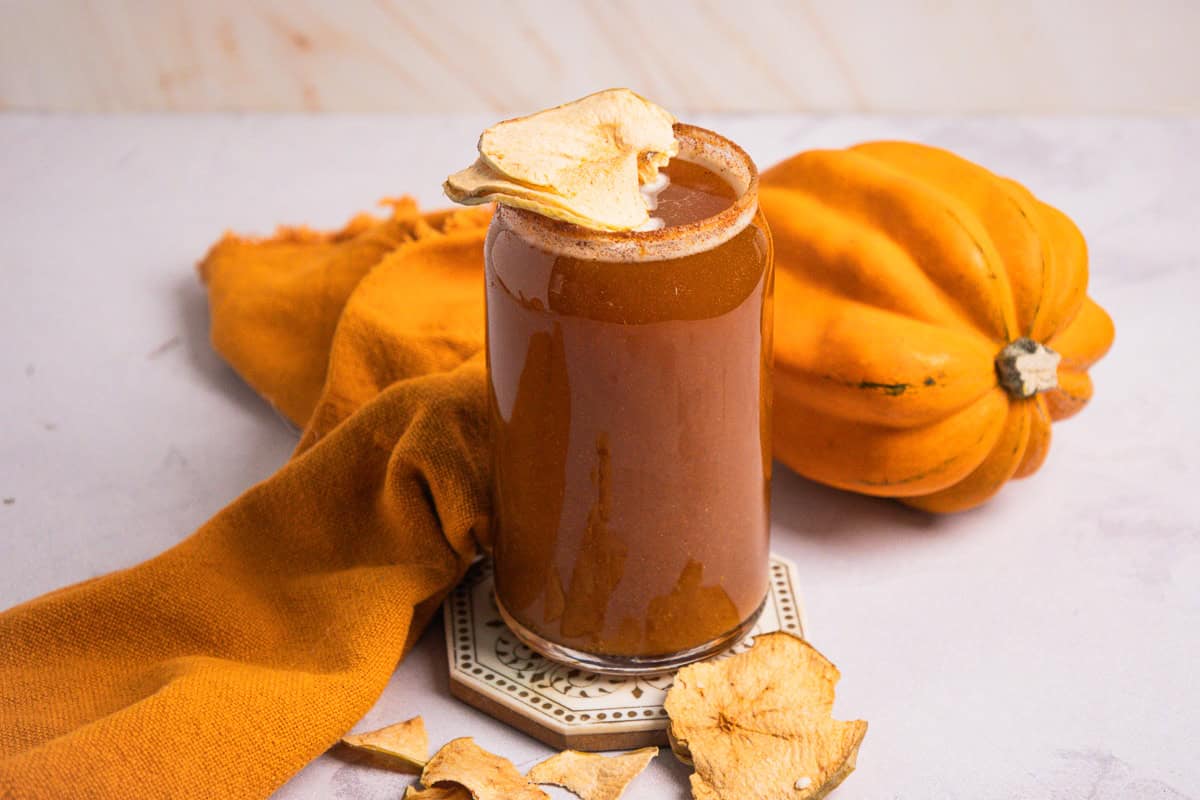 A glass of non-alcoholic pumpkin smoothie garnished with dried apple slices sits next to an orange squash and cloth, evoking the cozy flavors of pumpkin ale without the alcohol.