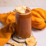 A glass of pumpkin smoothie topped with dried apple slices, next to a squash and orange cloth, evokes the cozy flavors of autumn-perfect for pairing with a chilled Pumpkin Ale or your favorite non-alcoholic beer.