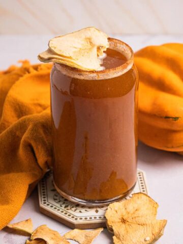 A glass of pumpkin smoothie topped with dried apple slices, next to a squash and orange cloth, evokes the cozy flavors of autumn-perfect for pairing with a chilled Pumpkin Ale or your favorite non-alcoholic beer.