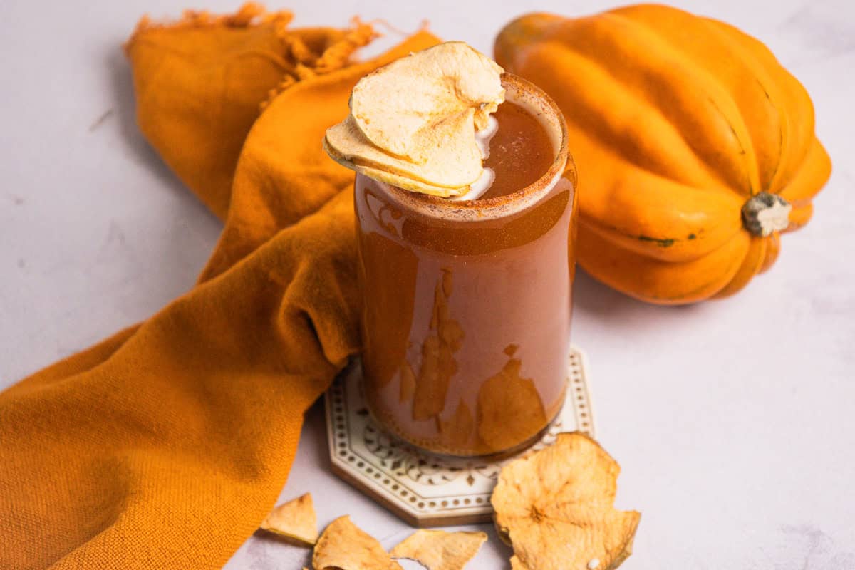 A tall glass of brown, non-alcoholic Pumpkin Ale garnished with dried apple slices, with a squash and orange cloth nearby.