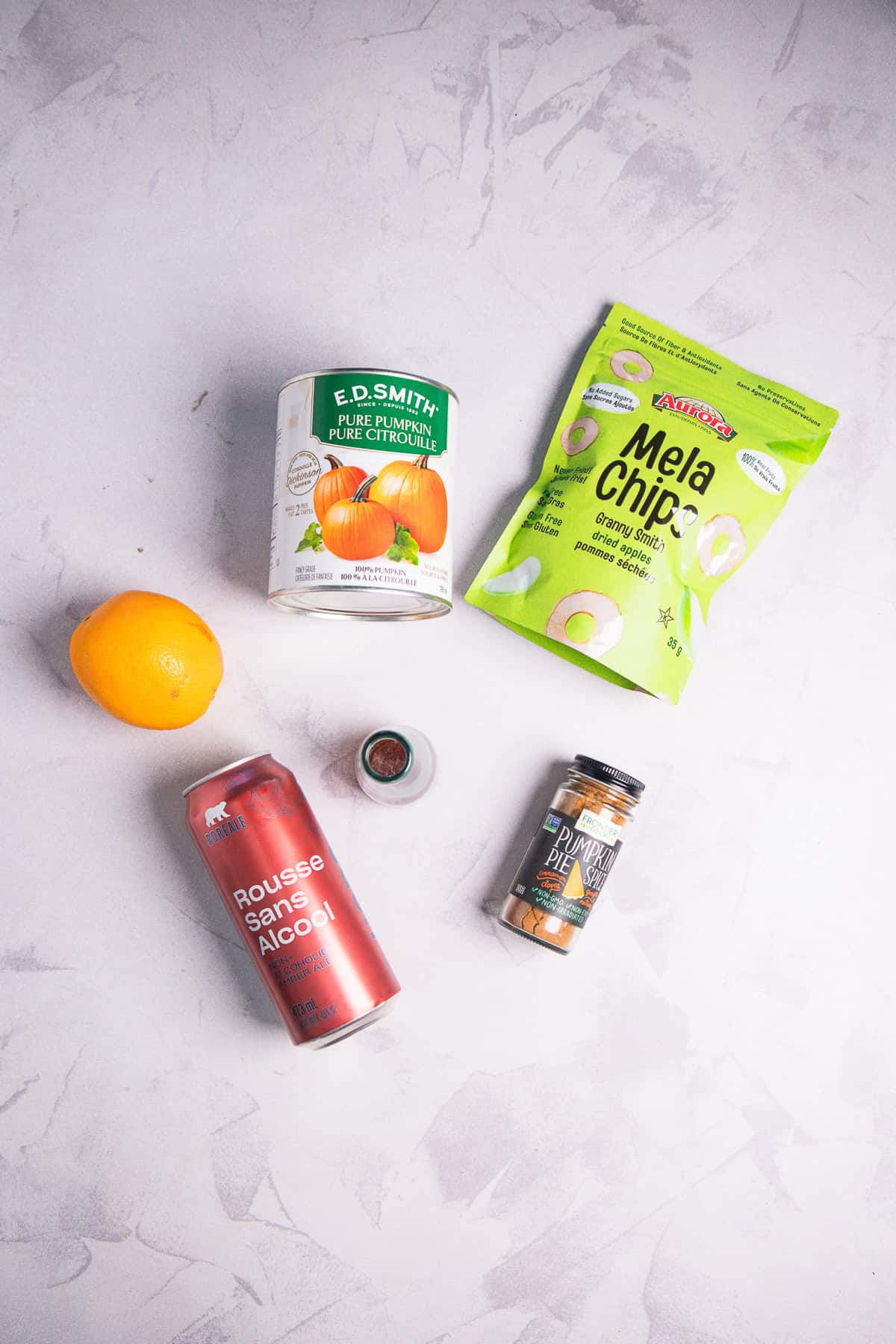 A lemon, pumpkin purรฉe can, apple chips bag, spice jar, spice container, and a refreshing can of Non-Alcoholic Beer are arranged on a light surface.