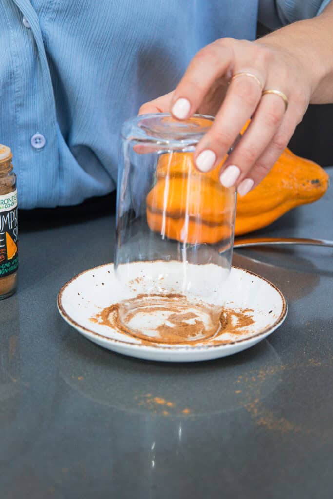 A hand dips a glass rim into a plate of brown spice, preparing it for a refreshing non-alcoholic pumpkin ale, with a jar and orange fruit nearby.
