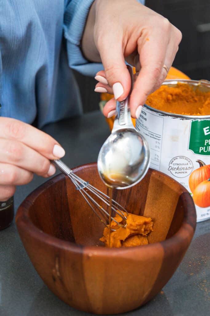 A person adds liquid, possibly non-alcoholic beer or pumpkin ale, to pumpkin puree in a wooden bowl with a whisk and an open can nearby.