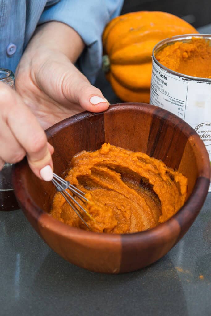 Hand whisking pumpkin puree in a wooden bowl, with a can of Non-Alcoholic Pumpkin Ale and a fresh pumpkin visible in the background.