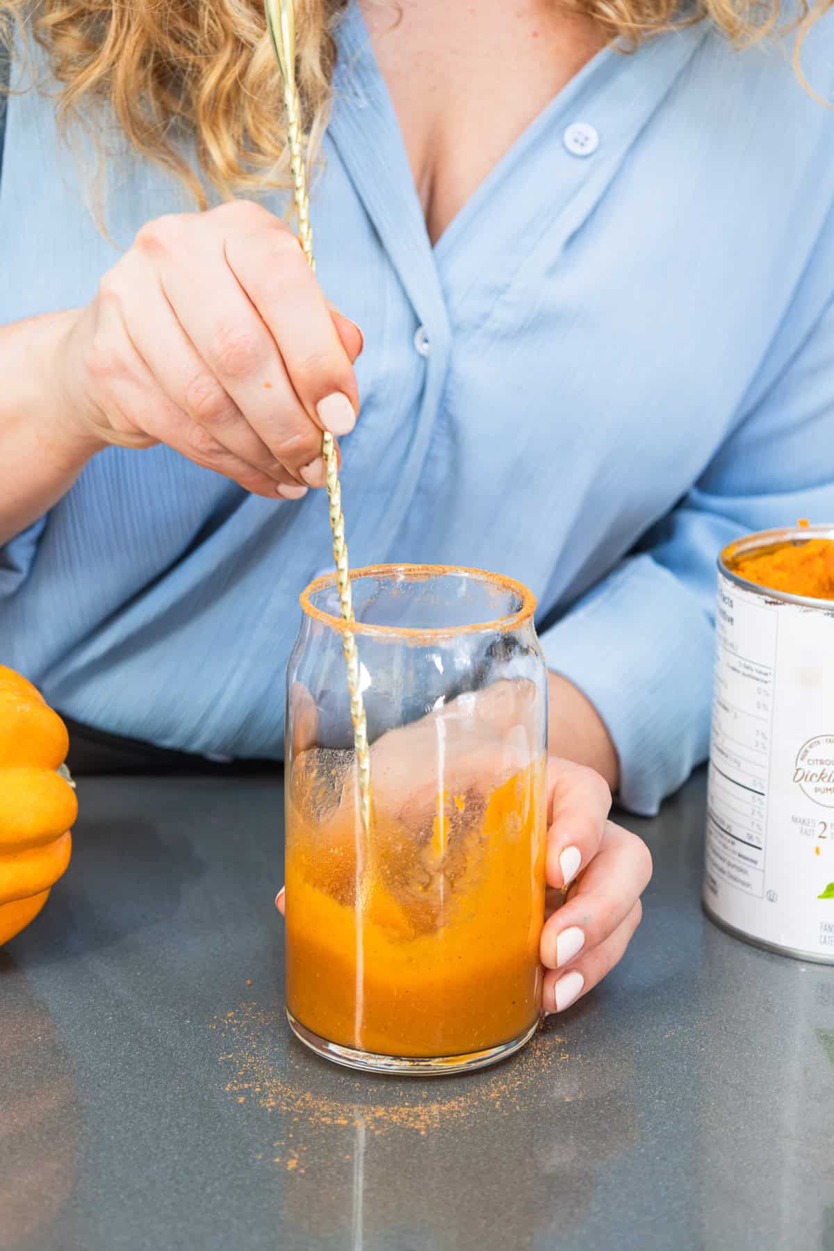 Person stirring an orange pumpkin drink in a glass with a gold spoon, wearing a blue shirtโevoking the cozy vibes of a festive Pumpkin Ale, but crafted as a delicious non-alcoholic treat.