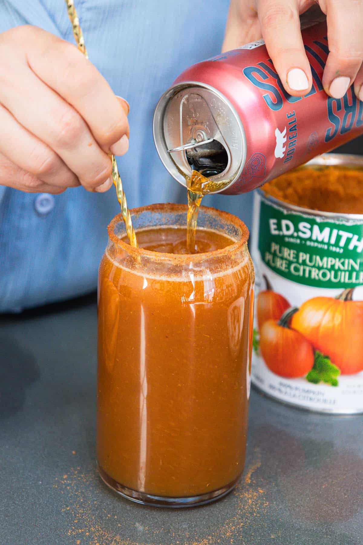 A hand pours non-alcoholic beer into a glass of pumpkin mixture beside an open can of pumpkin purรฉe.