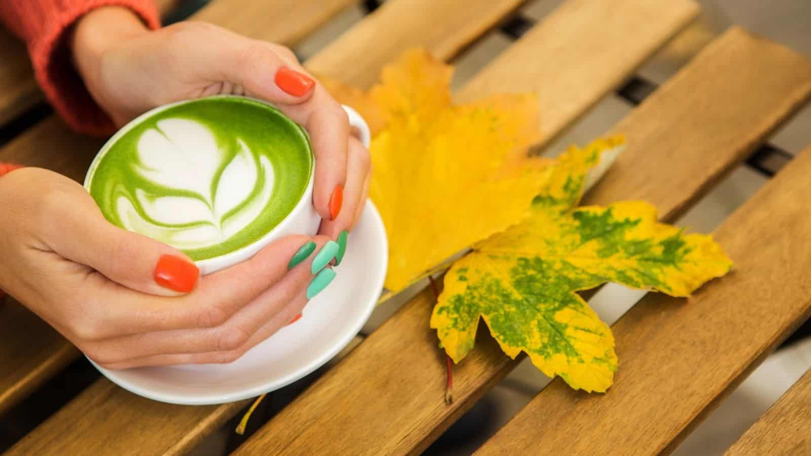 Hands with colorful nails hold a matcha latteโone of the top 7 coffee alternativesโon a wooden table with yellow autumn leaves nearby.
