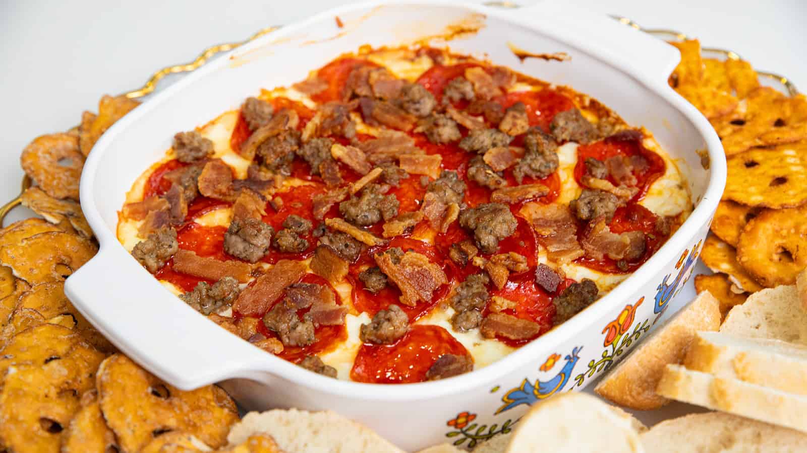 Baked dip topped with pepperoni, sausage, and bacon, surrounded by pretzels and bread slices.