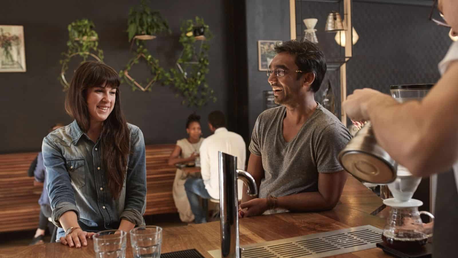 Two people laugh and chat at a coffee bar while a barista prepares a drink in a cozy cafรฉ, exploring 7 coffee alternatives on the menu.