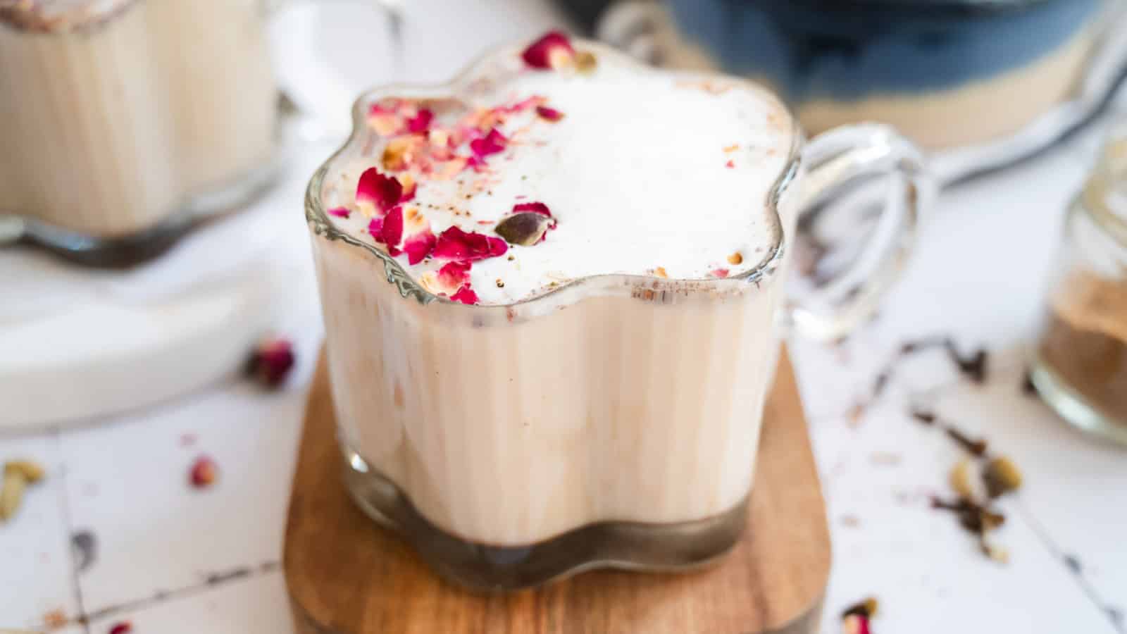 A glass mug of chai topped with rose petals sits on a wooden board, with honey and spices nearby.