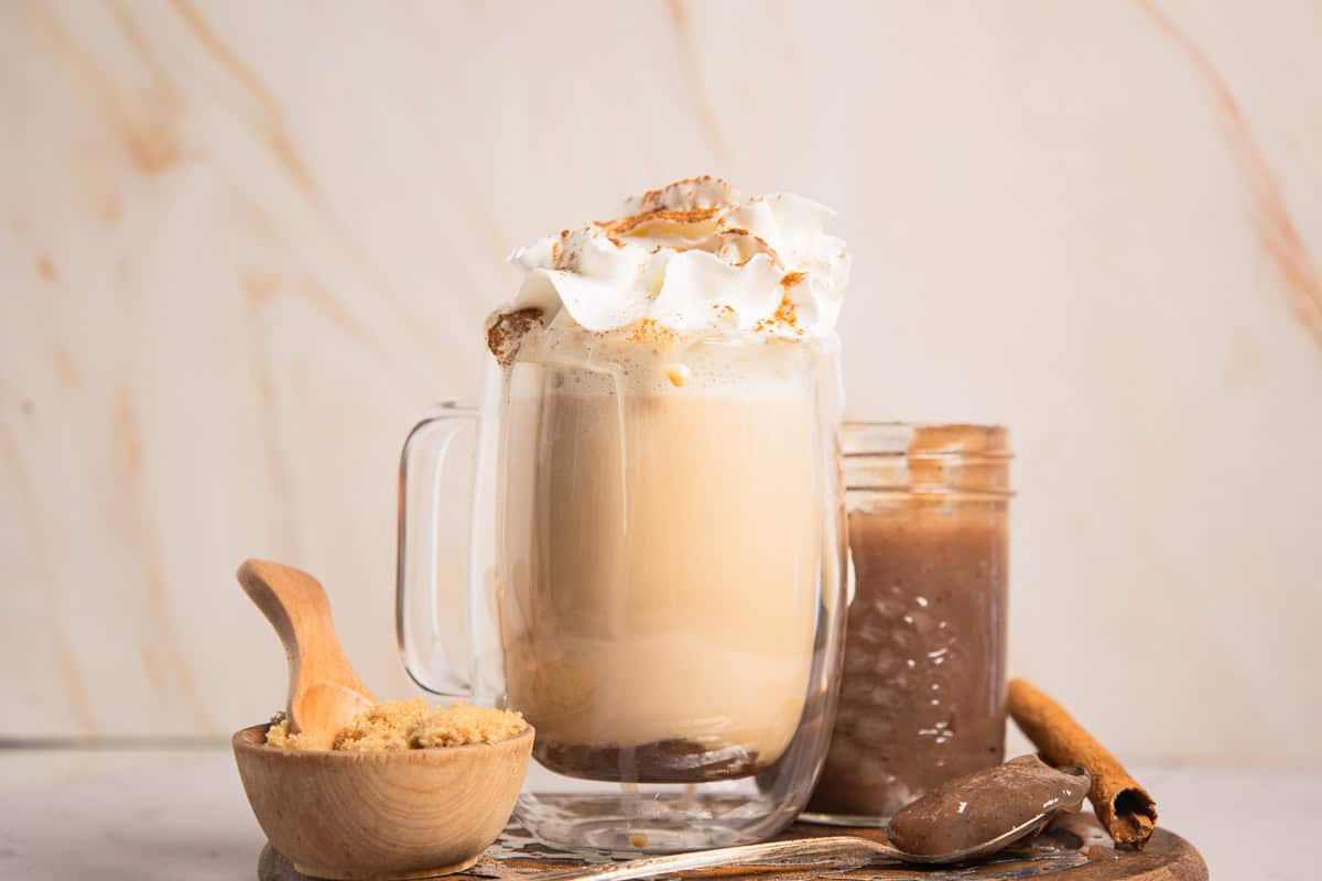 A glass mug of Banana Bread Latte topped with whipped cream, next to brown sugar and a jar of cinnamon spread.