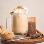 A glass mug of banana bread latte topped with whipped cream and cinnamon, served with brown sugar and a cinnamon stick.