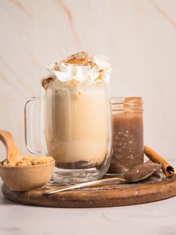 A glass mug of banana bread latte topped with whipped cream and cinnamon, served with brown sugar and a cinnamon stick.