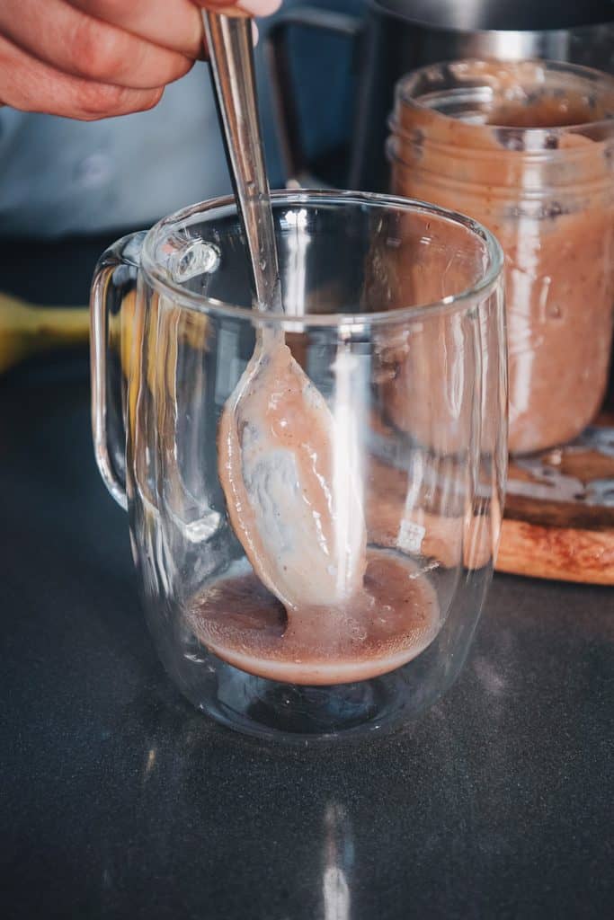 A metal spoon stirs a brown banana bread latte in a clear glass mug.
