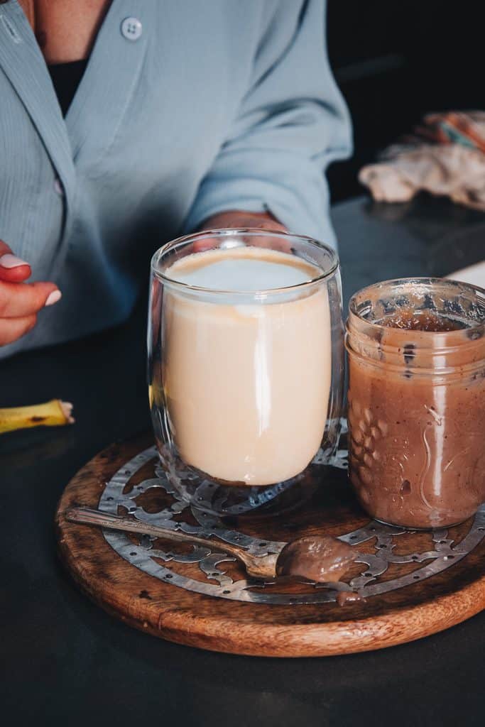 A glass of banana bread latte and a jar of nut butter on a decorative wooden tray.