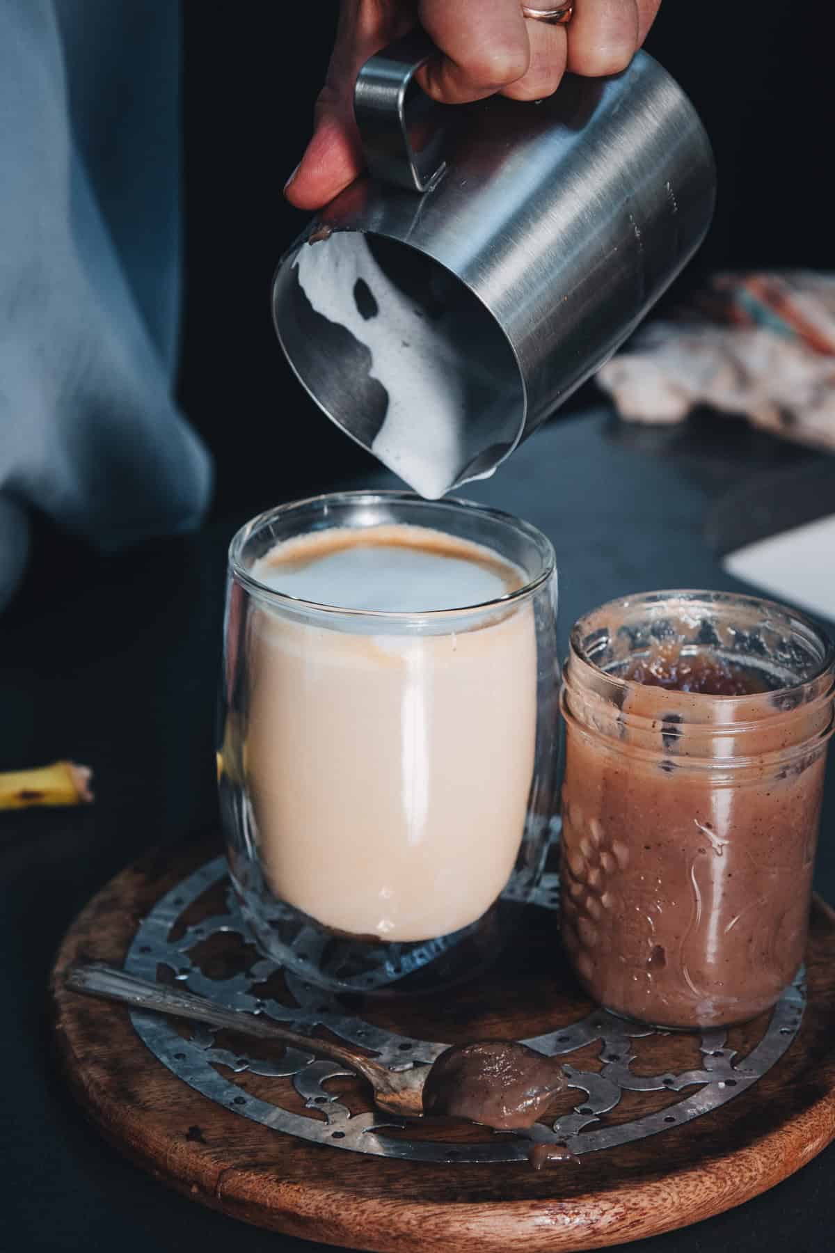 Steamed milk pours from a metal pitcher into a banana bread latte next to a jar of chocolate spread.