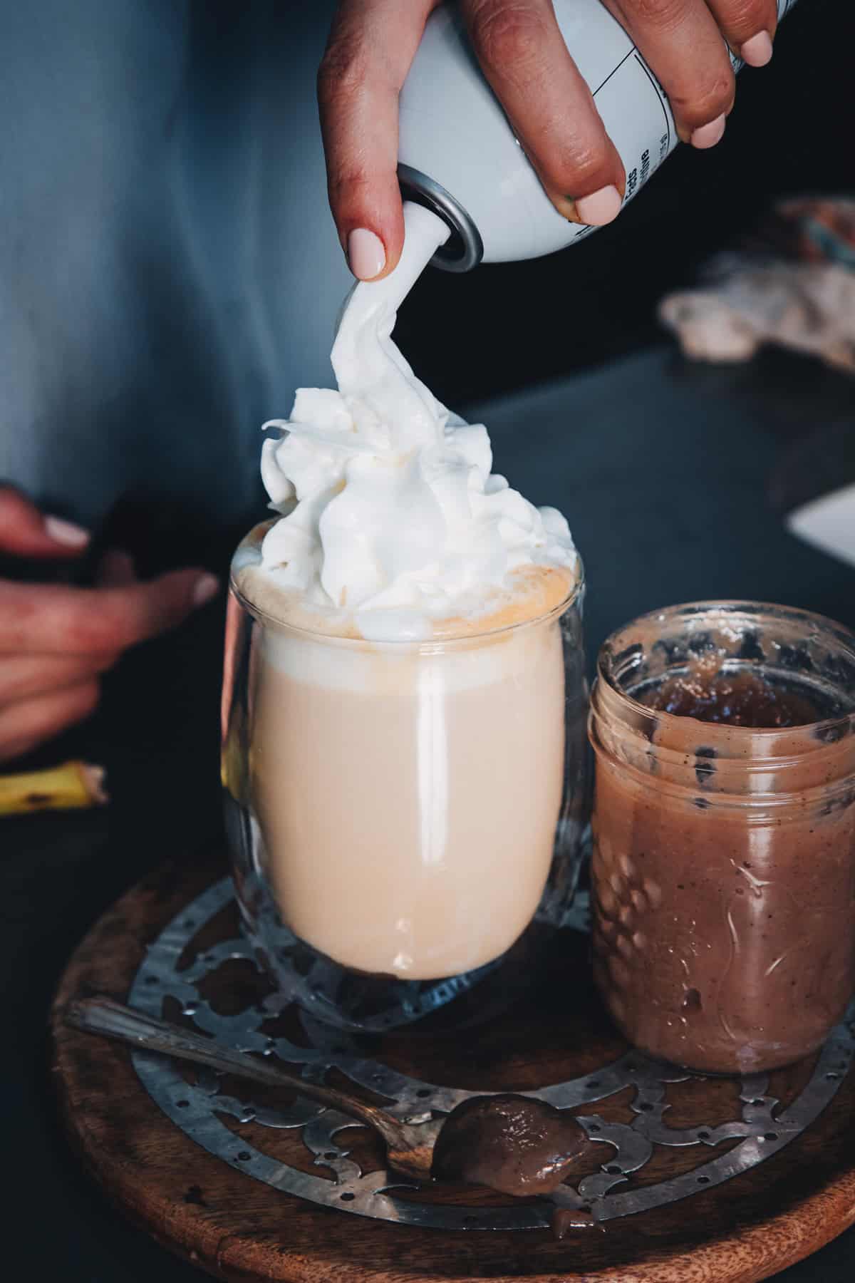 Hand dispensing whipped cream onto a banana bread latte, next to a jar of chocolate spread.
