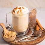 A glass mug of banana bread latte with whipped cream and cinnamon, served on a wooden board with brown sugar.