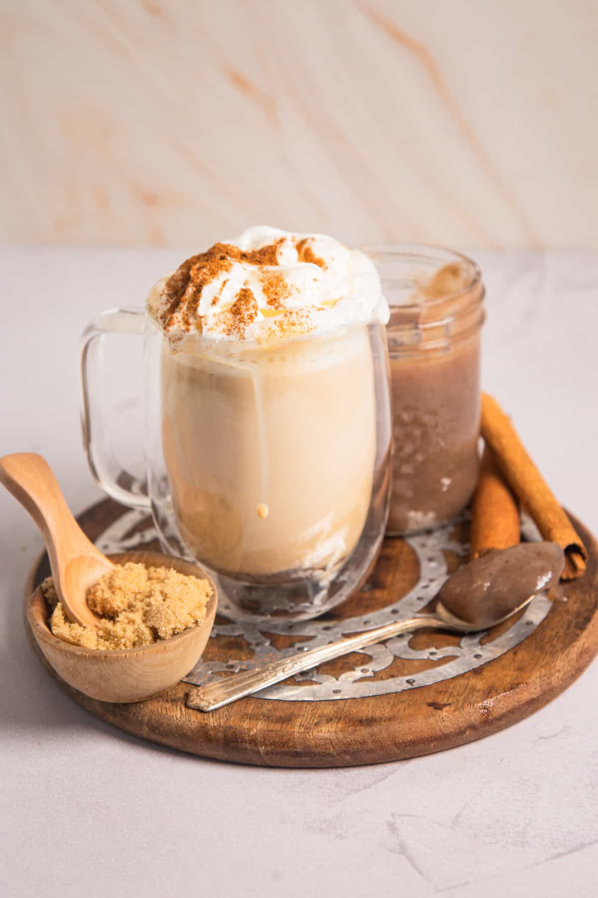 Glass mug of banana bread latte topped with whipped cream and cinnamon, served with brown sugar and cinnamon sticks.