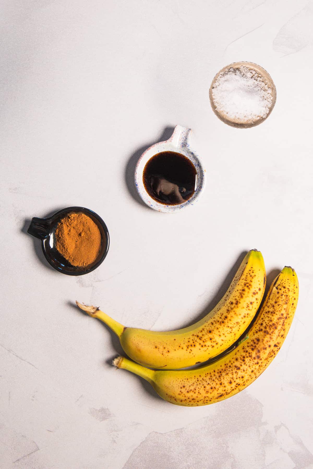 Two spotted bananas arranged with small bowls of cinnamon, dark syrup, and salt for a homemade banana coffee syrup.