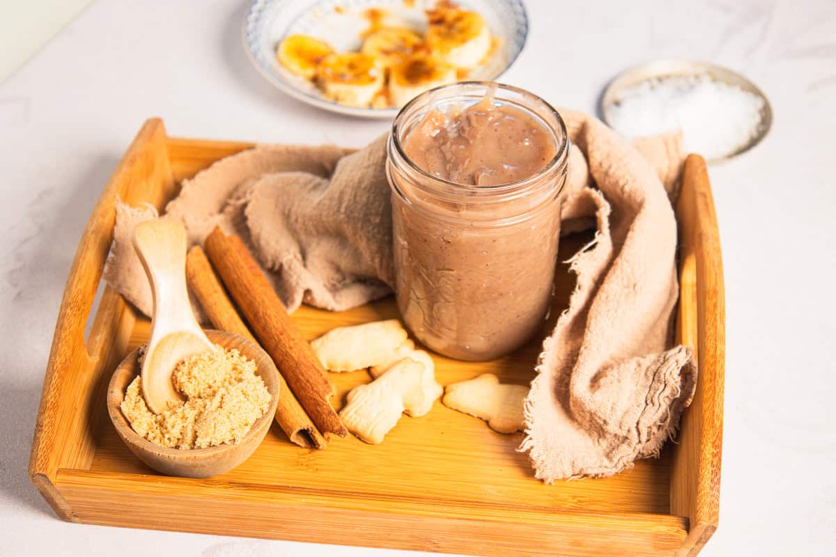 A jar of banana coffee syrup on a wooden tray with brown sugar, cinnamon sticks, and cookies.