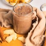 A glass jar of banana coffee syrup with small cookies, a cinnamon stick, and sliced bananas on a plate.