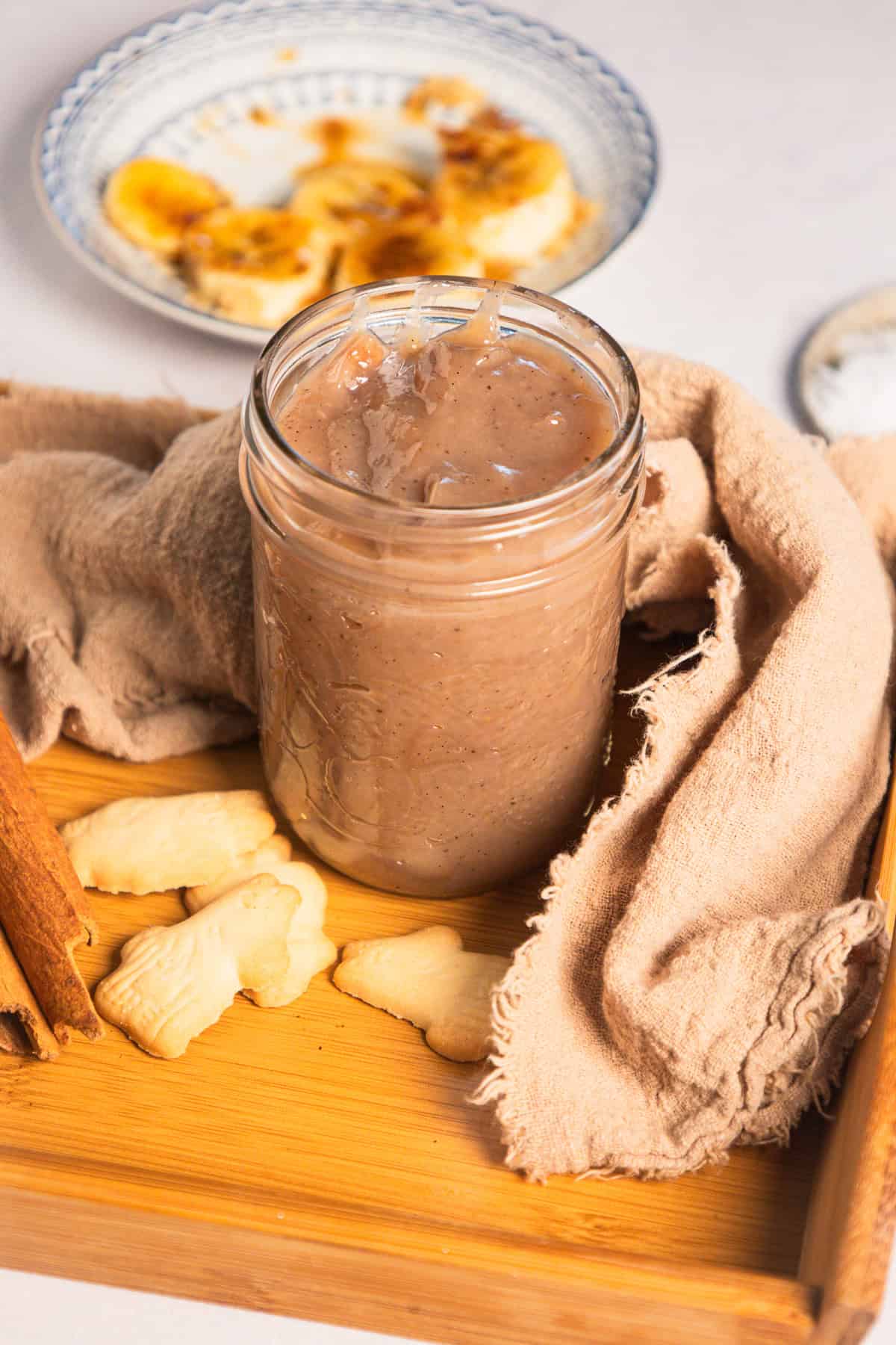 A glass jar of banana coffee syrup with small cookies, a cinnamon stick, and sliced bananas on a plate.