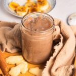 A glass jar of brown banana coffee syrup surrounded by cookies and a cinnamon stick on a wooden tray.