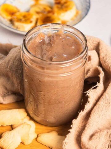 A glass jar of brown banana coffee syrup surrounded by cookies and a cinnamon stick on a wooden tray.