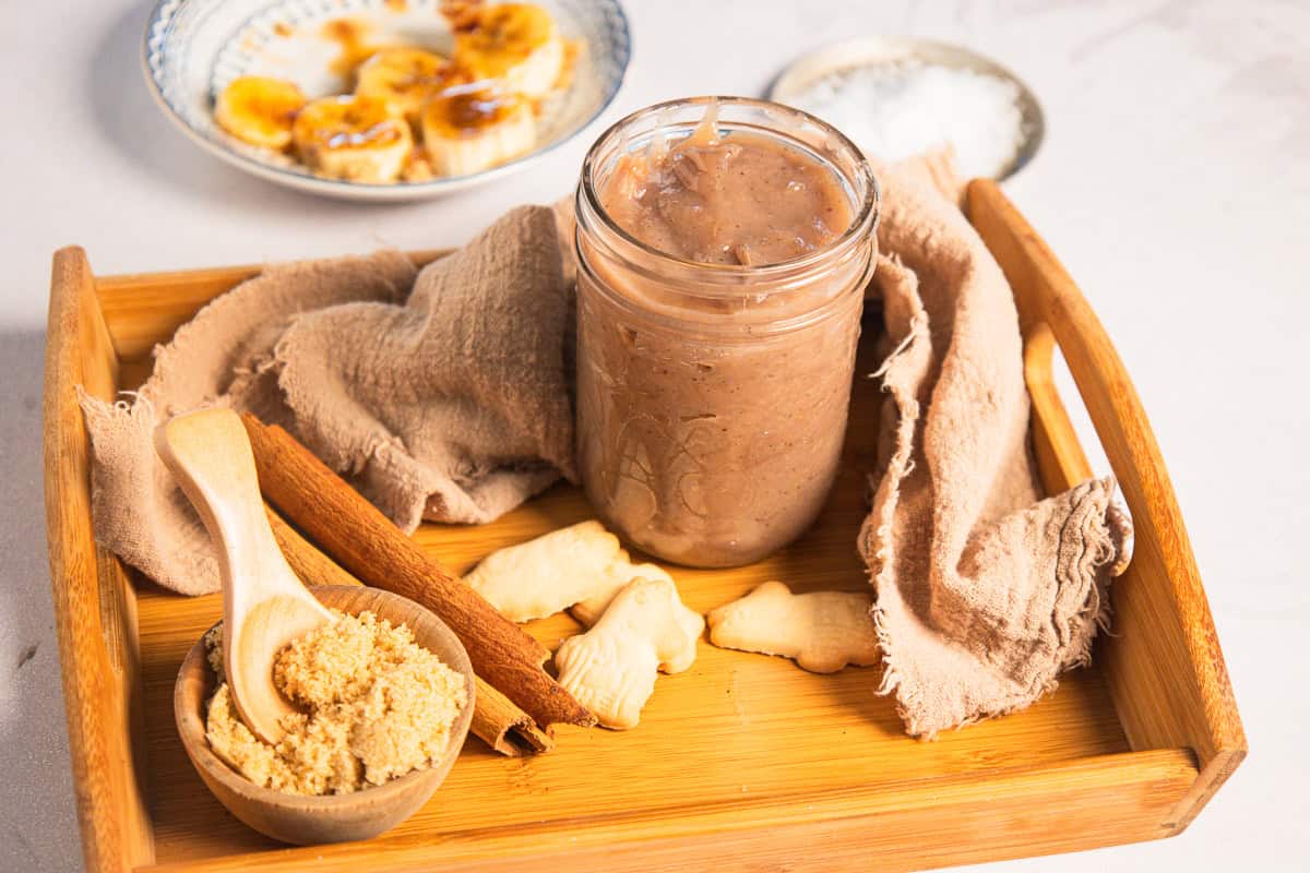 A glass jar of banana coffee syrup on a wooden tray with brown sugar, cinnamon sticks, and cookies.