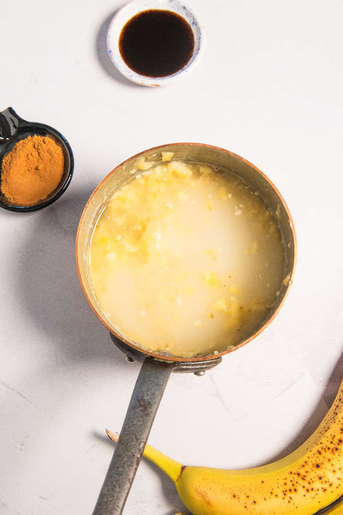 A small saucepan containing mashed bananas and liquid for a banana coffee syrup, with a bowl of cinnamon and a banana.
