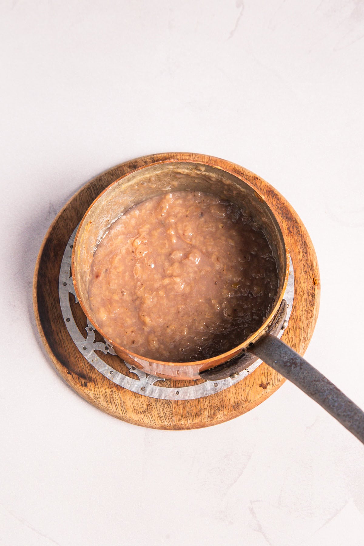 A copper pot of oatmeal drizzled with banana syrup sitting on a wooden trivet.