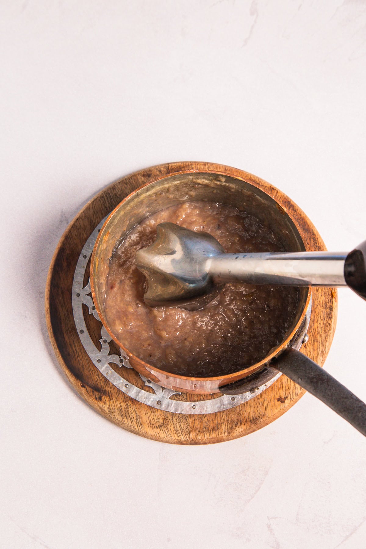 Immersion blender processing a thick banana syrup in a copper saucepan on a wooden trivet.